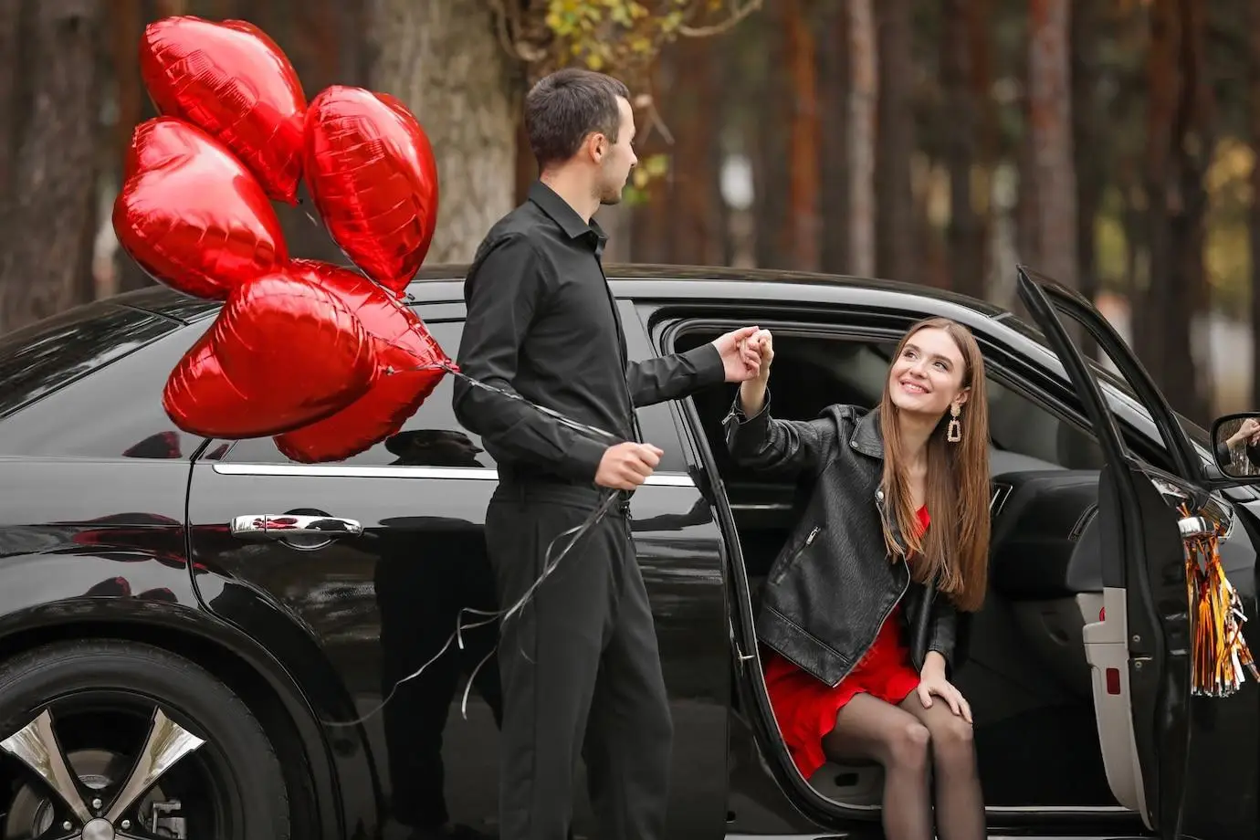 A man in black attire holds heart-shaped red balloons and helps a smiling woman in a red dress and black jacket step out of a black luxury car in a wooded area.