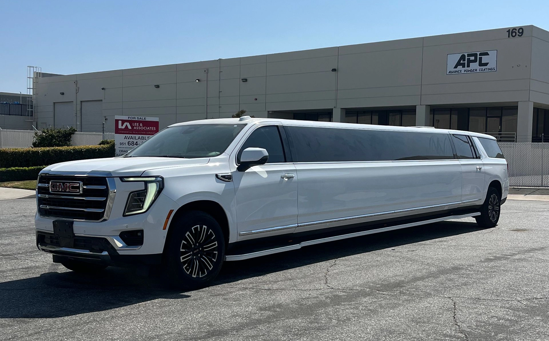 White 2025 GMC Yukon Stretch Limousine parked in front of an industrial building under a clear blue sky.