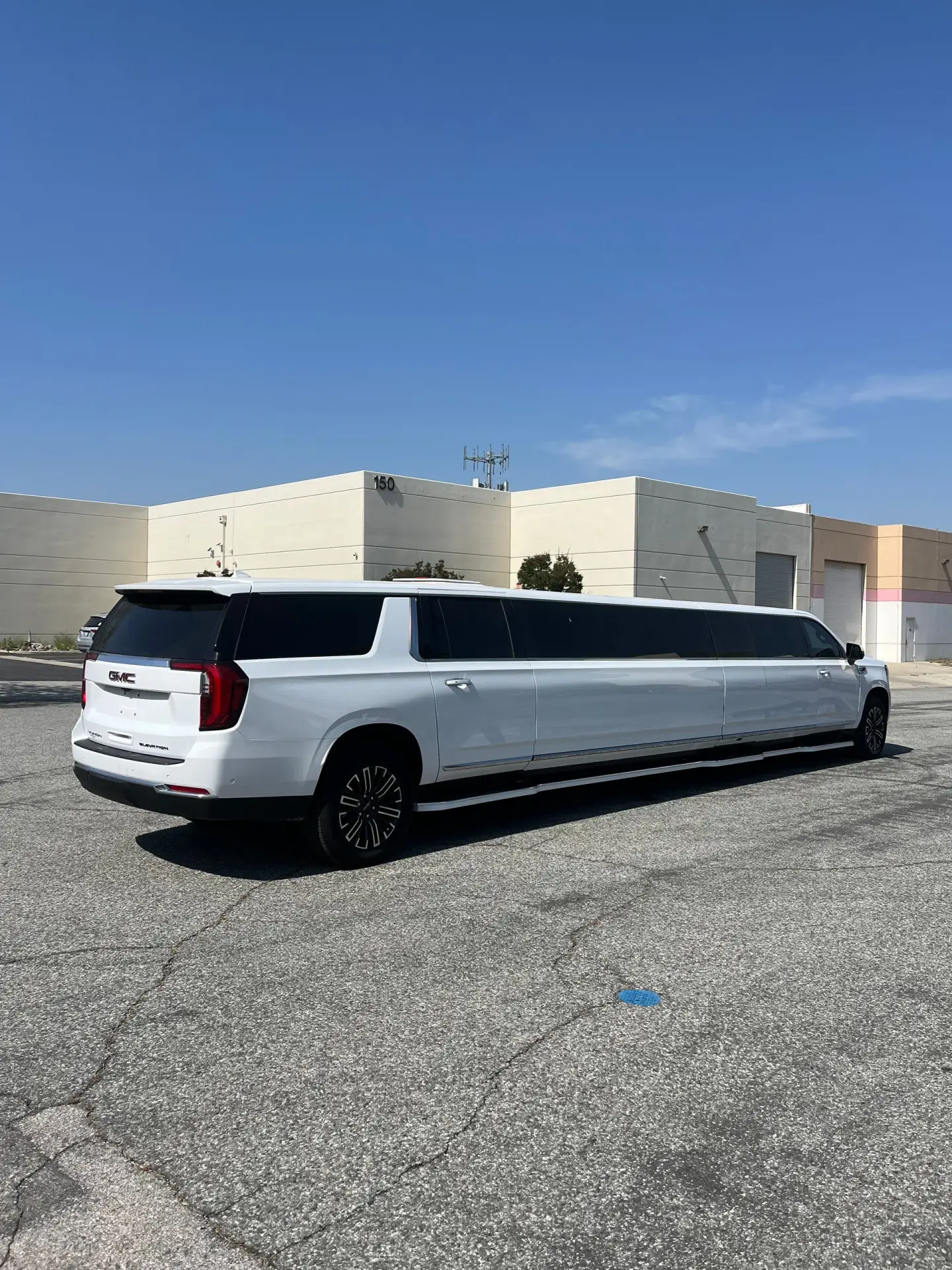 Rear-side view of a white 2025 GMC Yukon Stretch Limousine by Basking Ridge Limousine, parked outside a modern commercial facility.