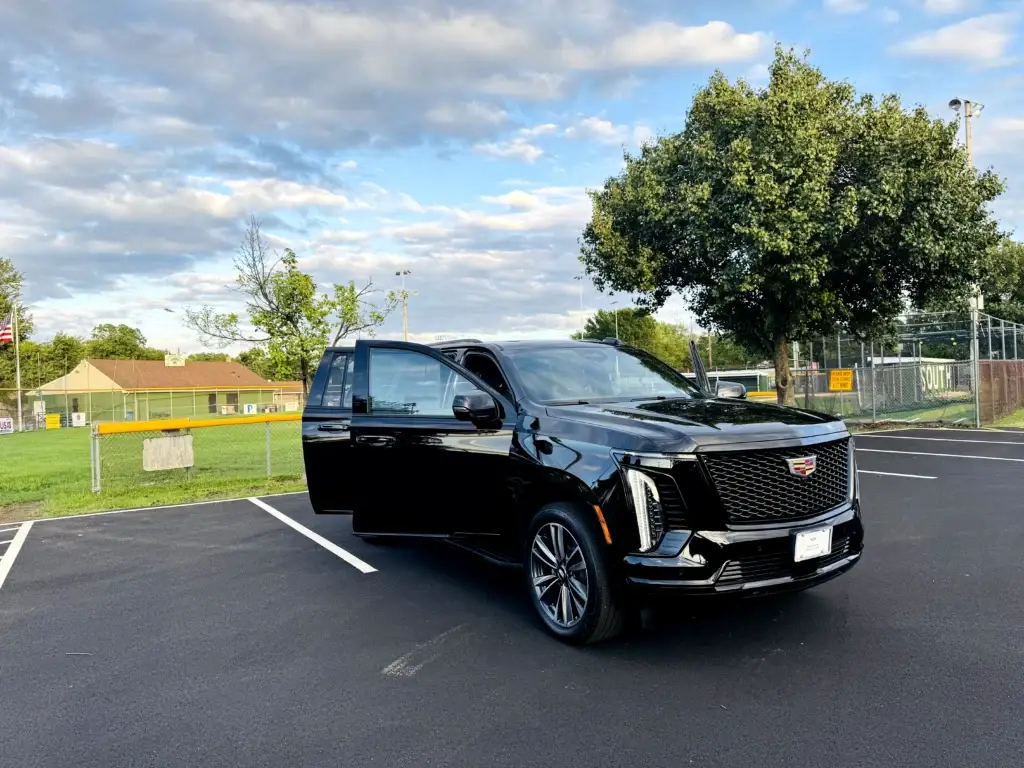A black 2025 Cadillac Escalade Platinum SUV is parked in an empty lot on a clear afternoon, with its driver’s door open. Behind it are a baseball field, trees, and a partly cloudy blue sky.