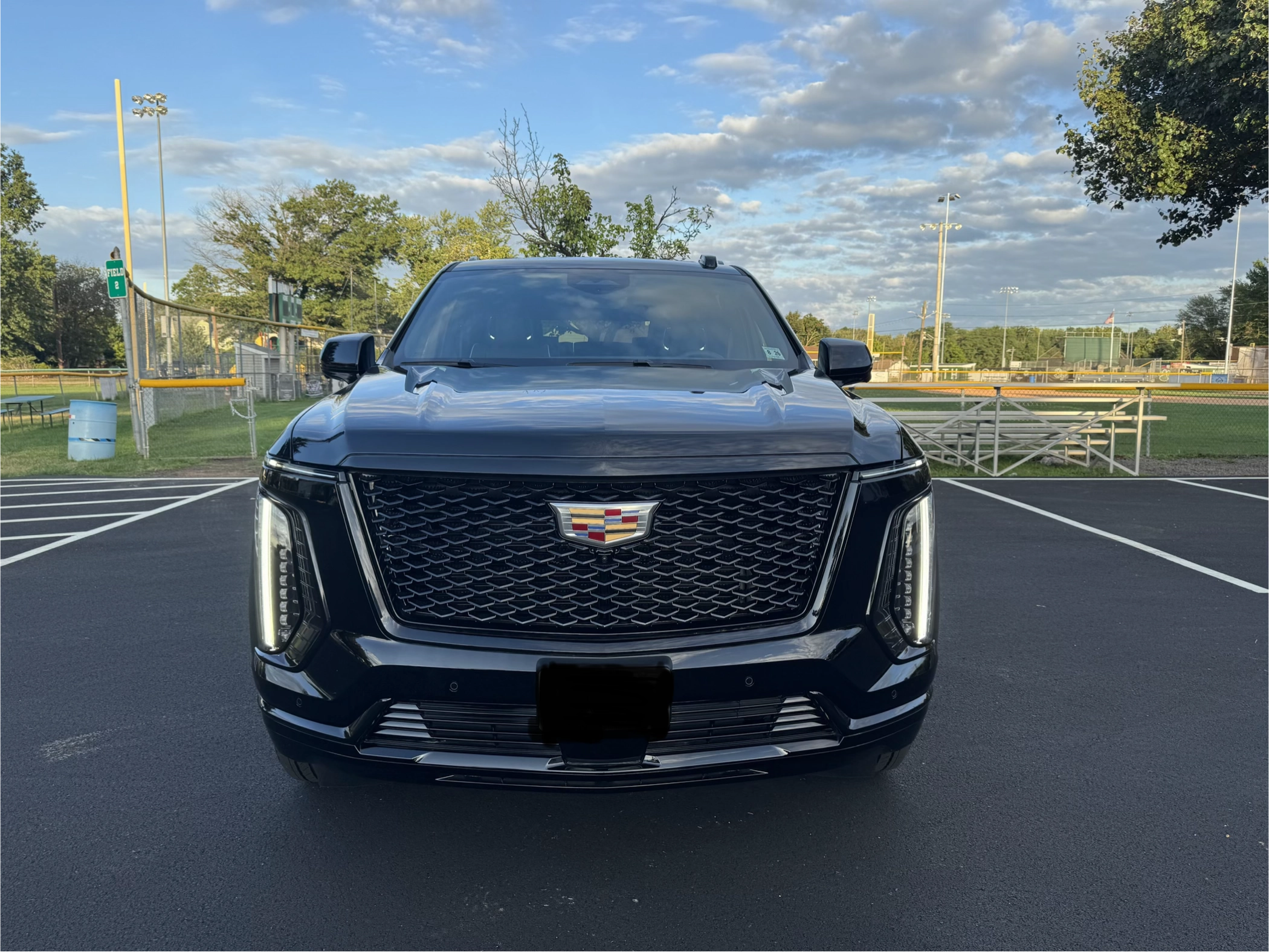 Luxury black 2025 Cadillac Escalade Platinum SUV front view with both doors open in a parking lot near a baseball field under blue sky – Basking Ridge Limousine Services, New Jersey.