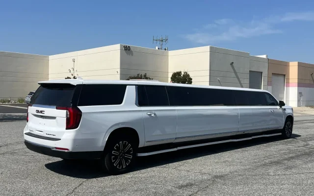 Rear-side view of a white 2025 GMC Yukon Stretch Limousine by Basking Ridge Limousine, parked outside a modern commercial facility.