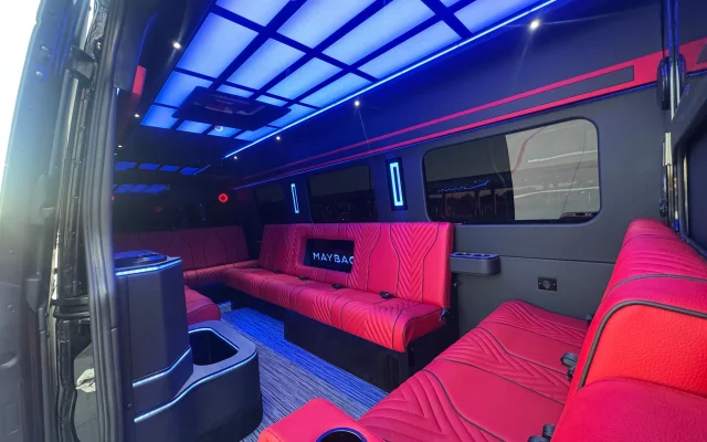 Interior of the Maybach Sprinter party bus lit in blue and magenta, with red quilted lounge benches, moulded cup wells and a console cabinet beside the door