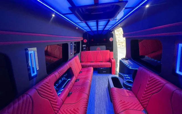 View from the rear of the Sprinter party bus toward the open side door, showing facing red leather benches, cabin speakers and blue illuminated window strips